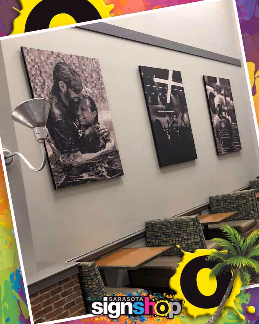 Three black-and-white photos hang on a wall above patterned chairs and tables in a cafe. The image has a colorful border with the Sarasota sign shop logo and palm trees at the bottom.