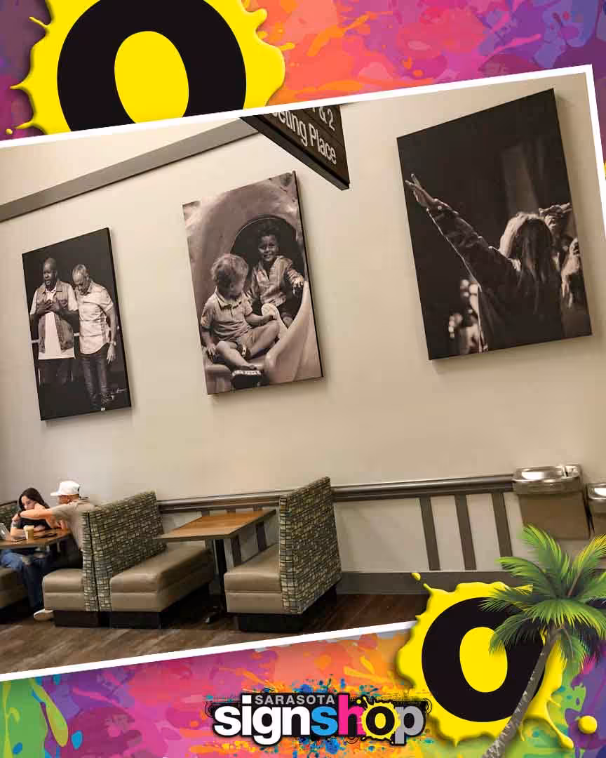 Three black-and-white photos hang above two booth tables in a restaurant. Two people sit at one table. The image has a colorful border with Sarasota Sign Shop text at the bottom.