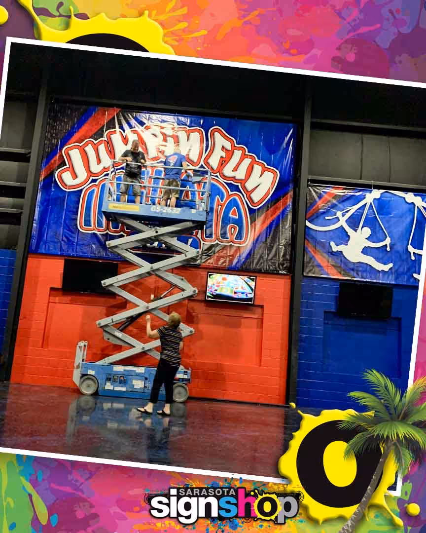 Two people on a lift install a large Jumpin Fun banner on a red and blue wall inside a gym. A third person stands below, guiding them. The Sarasota Sign Shop logo is at the bottom.