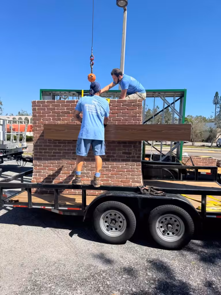 Two men in blue shirts from a Real Estate Sign Company work on a fake brick wall being lifted onto a trailer by a crane, assembling or securing it under a clear blue sky in an outdoor area.