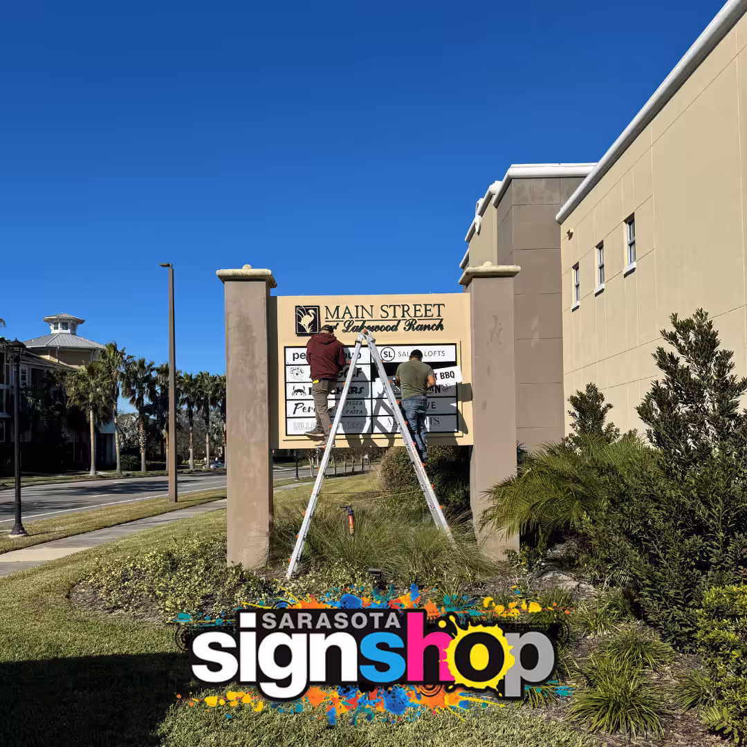 Two people on ladders update a directory sign outside a building under a clear blue sky. Sarasota Sign Shop logo is at the bottom of the image.