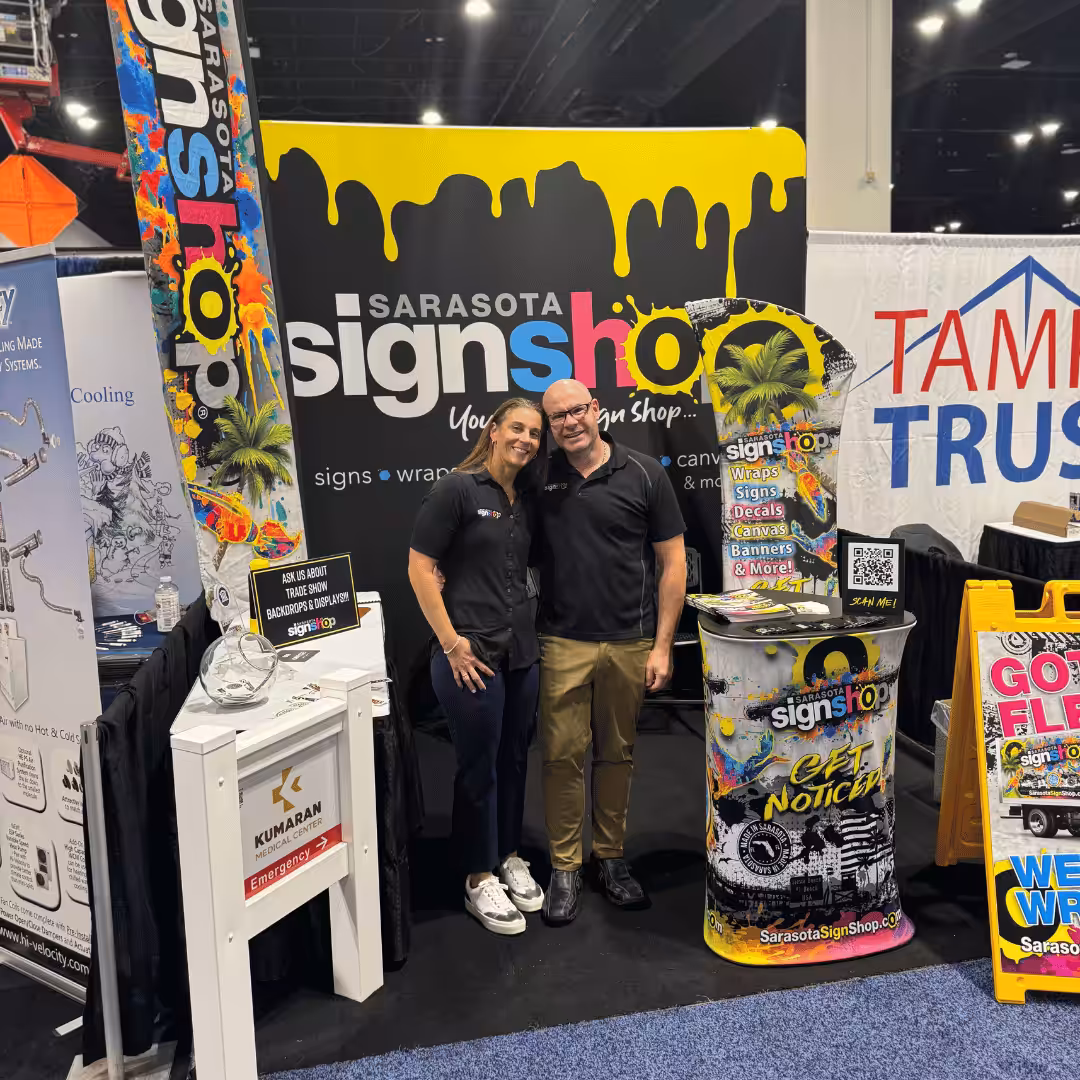 Two people stand smiling in front of a Sarasota Sign Shop display booth at a trade show, surrounded by colorful signs, banners, and promotional materials.