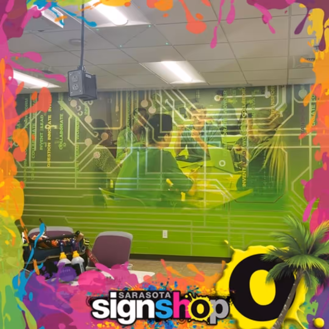 People working at a table with laptops in a green tech-themed room, featuring office wall murals with a circuit board design and a colorful splatter border reading Sarasota Sign Shop.