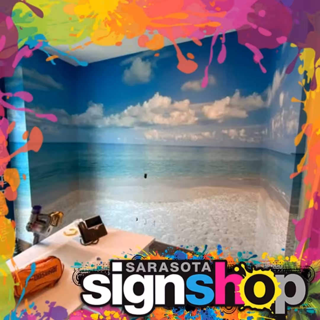 A small office features a beach and ocean wall mural under a blue sky. Colorful paint splashes border the scene, highlighting Sarasota Sign Shop’s custom wallcoverings Sarasota at the bottom.