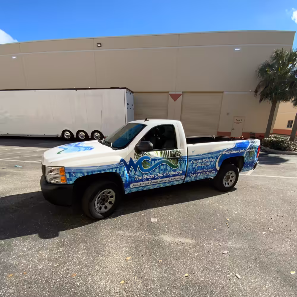 A white pickup truck with blue and green water-themed graphics and The Water Club of America, expertly wrapped by Sarasota Sign Shop, is parked outside a beige industrial building on a sunny day.