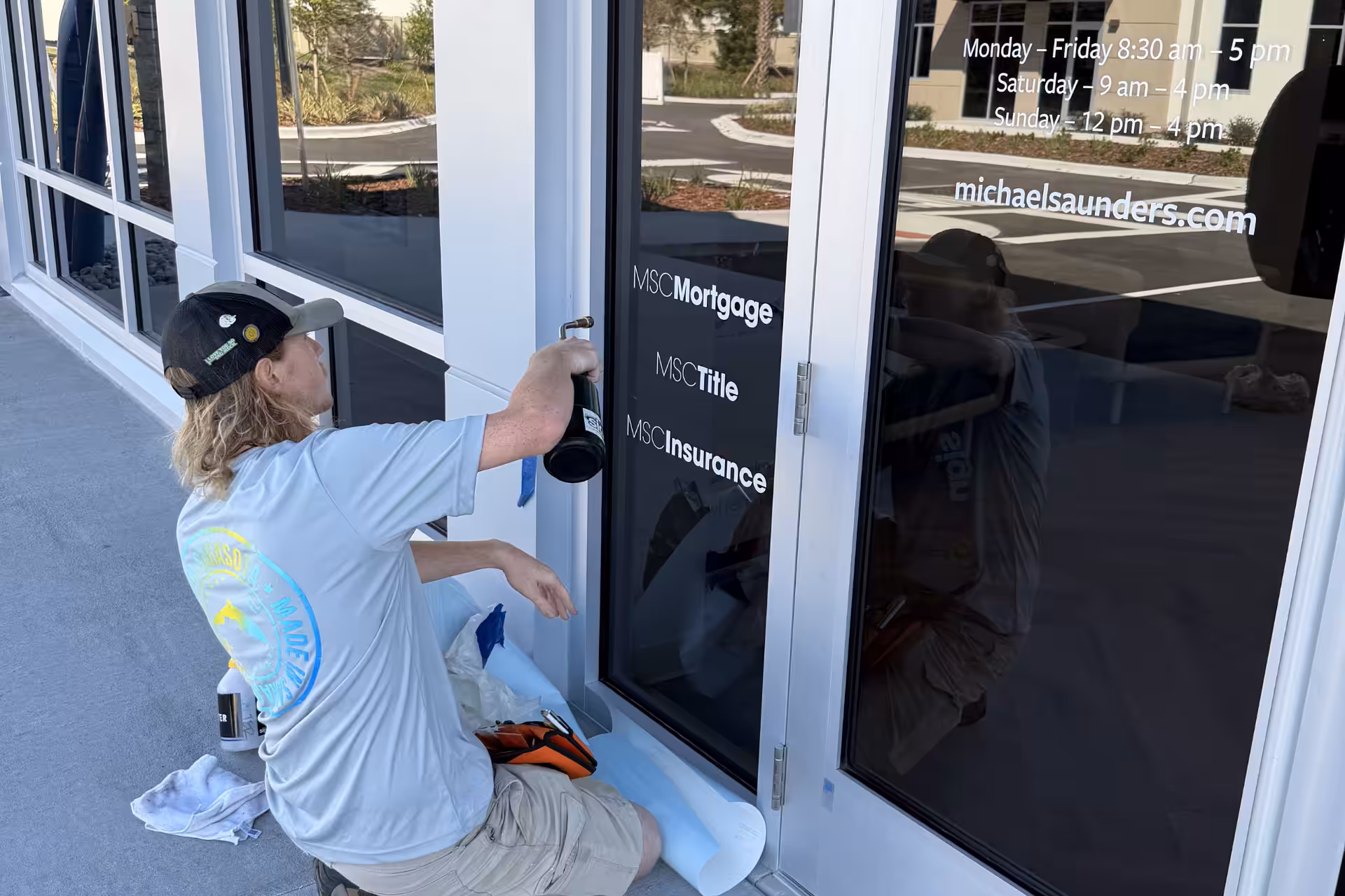 A person kneeling outside a glass door in Sarasota applies white vinyl lettering listing business services such as mortgage, title, and insurance. Hours of operation are also displayed on the glass.