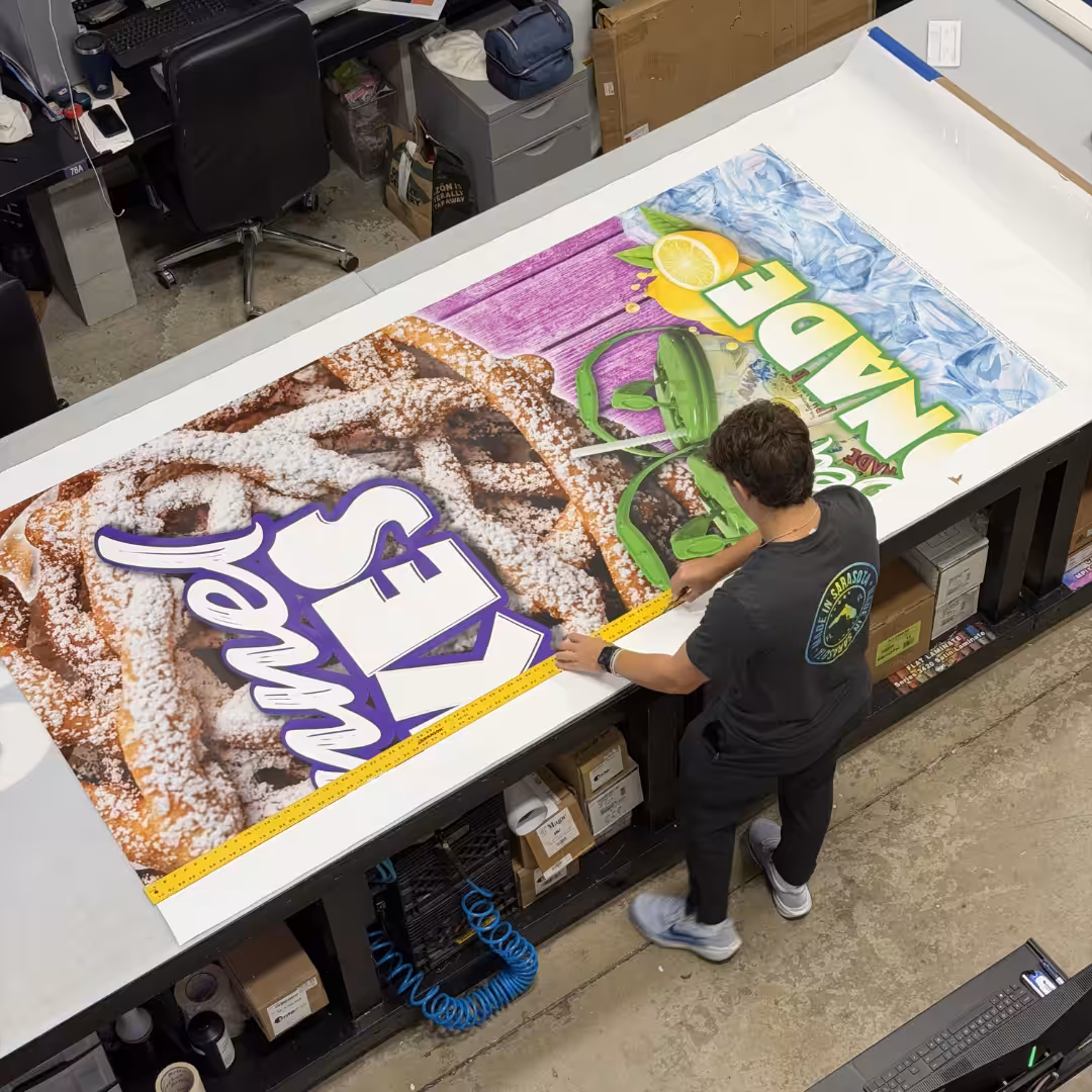 A person stands at a large table in a Venice print shop, examining two colorful printed banners—one featuring funnel cakes and the other lemonade—amid scattered supplies and equipment.