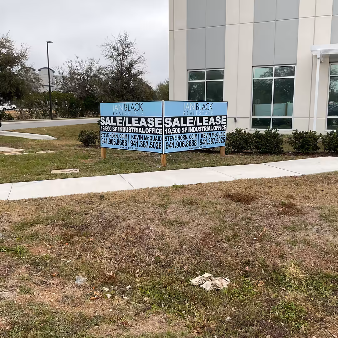 Two blue and white real estate For Sale/Lease signs stand in front of a modern building with large windows, offering industrial/office spaces. The grassy area around features some patches of dirt.