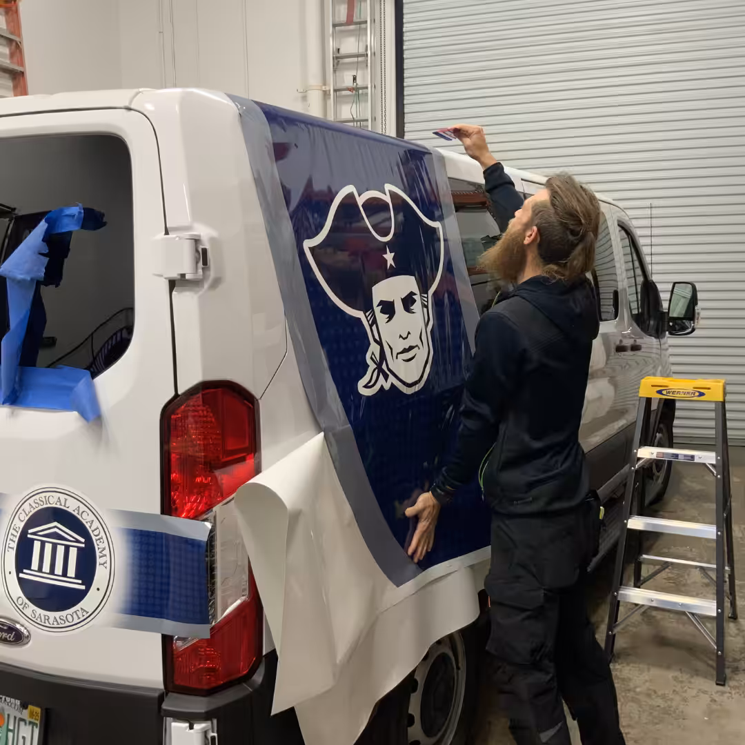 A man applies a large blue decal featuring a patriot head logo, often seen in schools & sports, to the side of a white van inside a garage, with a stepladder and tools nearby.