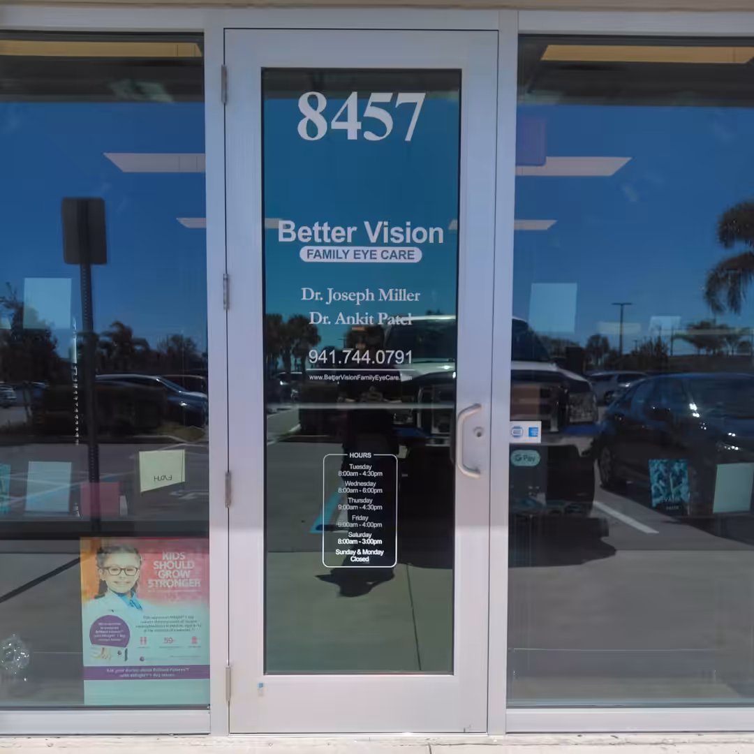 Glass door of Better Vision Family Eye Care, a healthcare clinic, displays doctors’ names, contact info, and hours. Reflections of cars and palm trees shimmer in the glass.