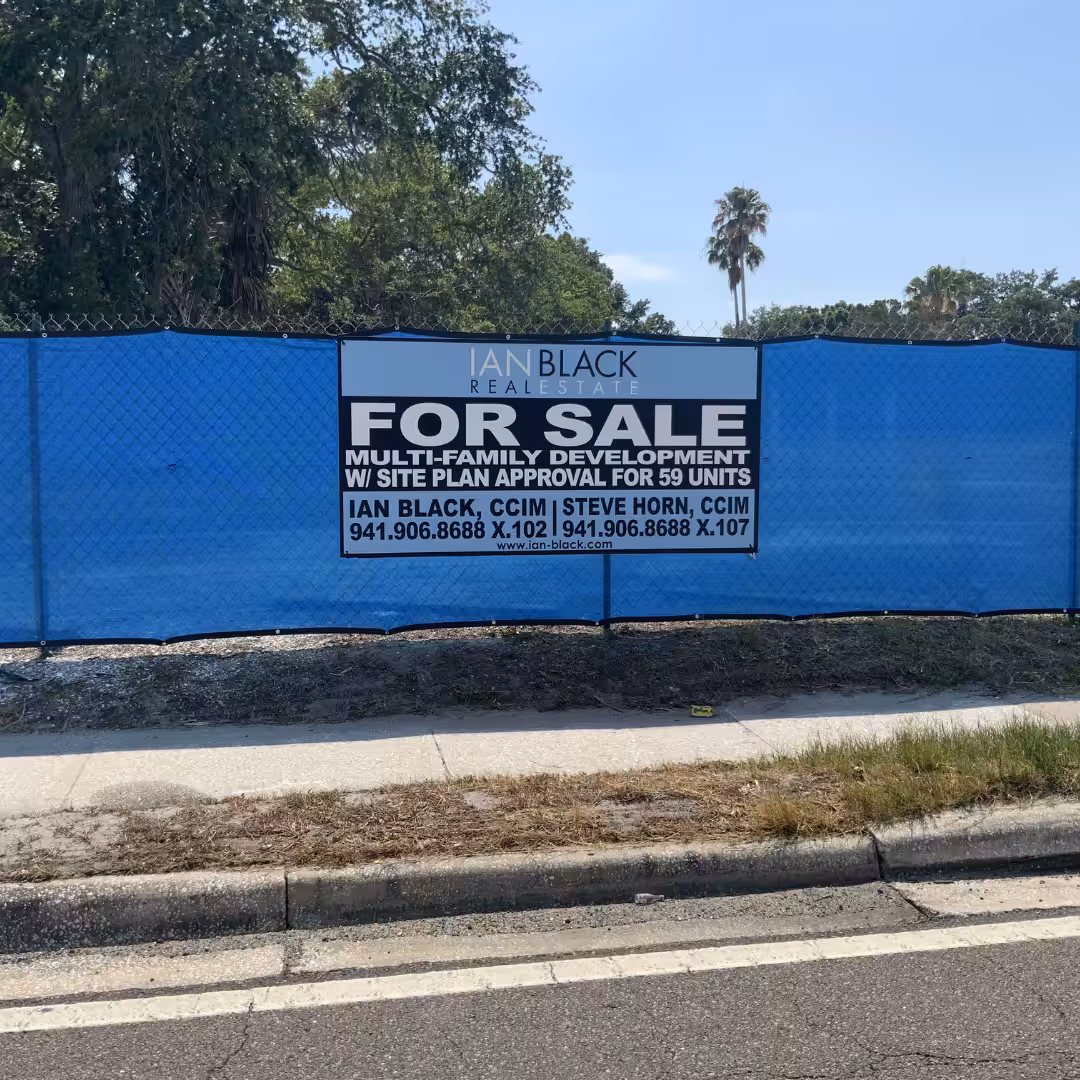 A blue fence by a roadside features banners and a For Sale sign for a multi-family development with site plan approval for 59 units. The sign lists Ian Black Real Estate agents’ contact info, with trees behind the fence.