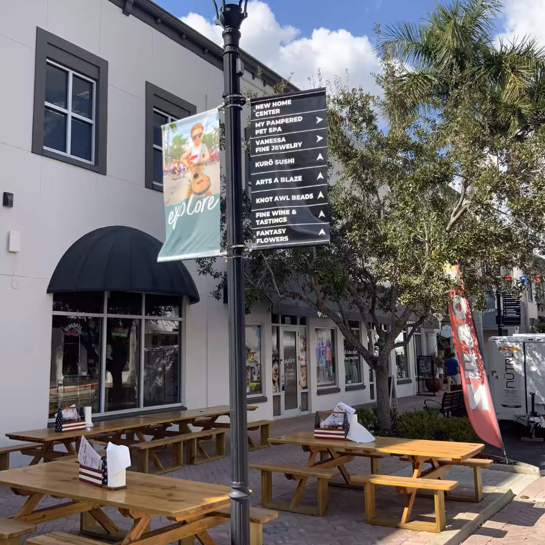 Outdoor dining area with wooden picnic tables, retail directional sign listing businesses, American flag-themed napkin holders, and a tall palm tree beside a two-story building under a partly cloudy sky.