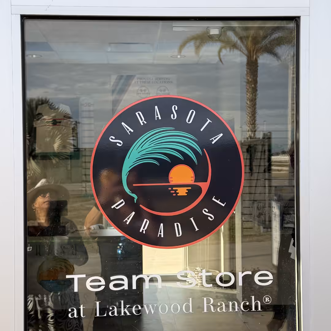 Glass door with a round Sarasota Paradise logo featuring a palm tree and sunset. Below, text reads Team store at Lakewood Ranch for Schools & Sports. Reflections of a palm tree and people are visible in the glass.