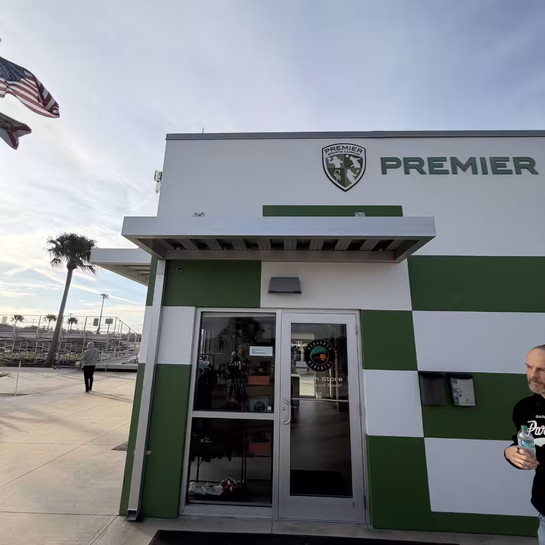 A man stands outside a white and green Premier sports building, with glass doors, an American flag, palm trees, and nearby schools & sports facilities visible in the background.