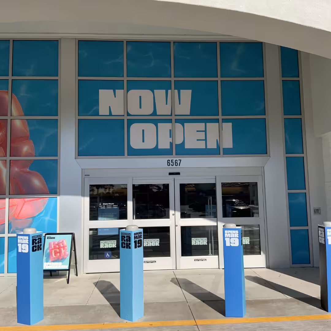 The entrance of a retail store features large blue signs reading NOW OPEN above the doors. Four blue posts and a sandwich board sign sit out front, with address number 6567 displayed above the entrance.
