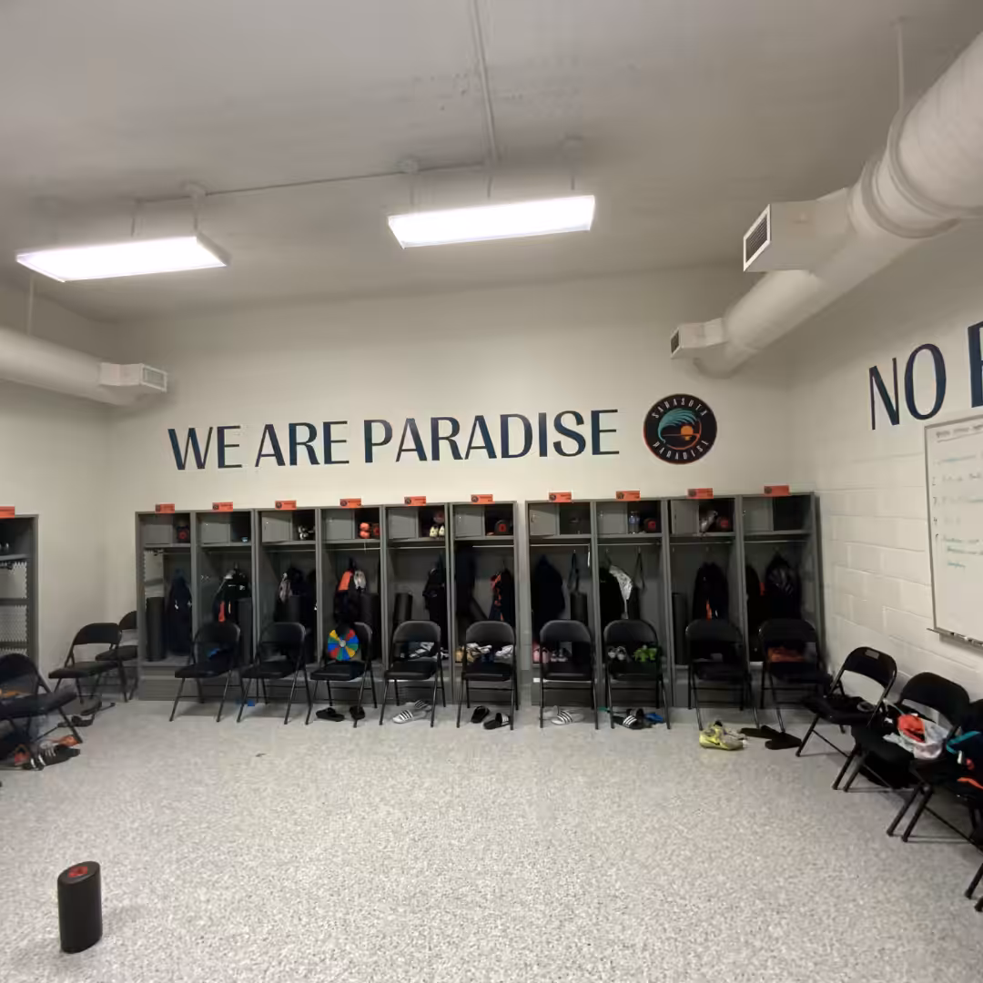 A locker room with open lockers, sports gear, and chairs—perfect for schools & sports. WE ARE PARADISE is written above the lockers. The floor is mostly clear, with a few scattered items.