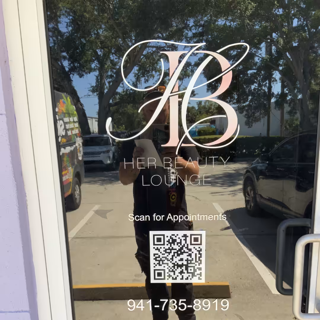 Glass door of this retail space features the Her Beauty Lounge logo, a QR code for appointments, and a phone number (941-735-8919). Cars and trees are reflected in the glass.