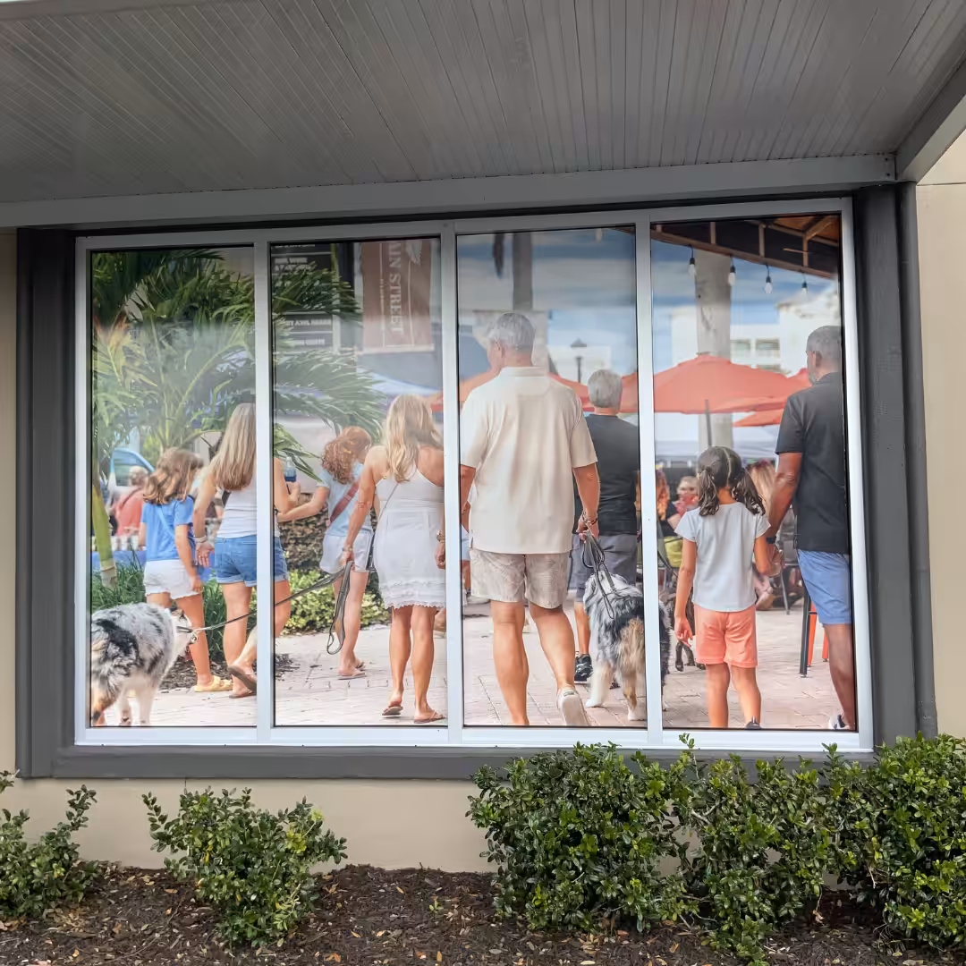 A retail window display features a large photo of people walking outdoors near shops and restaurants, with two dogs and several adults and children. Green bushes are planted below the window.
