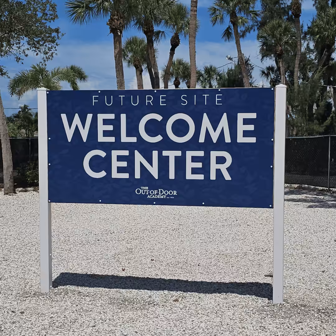 A blue sign reads Future Site Welcome Center with The Outdoor Academy logo below, standing on gravel with palm trees and a fence—highlighting an exciting project for builders & developers.