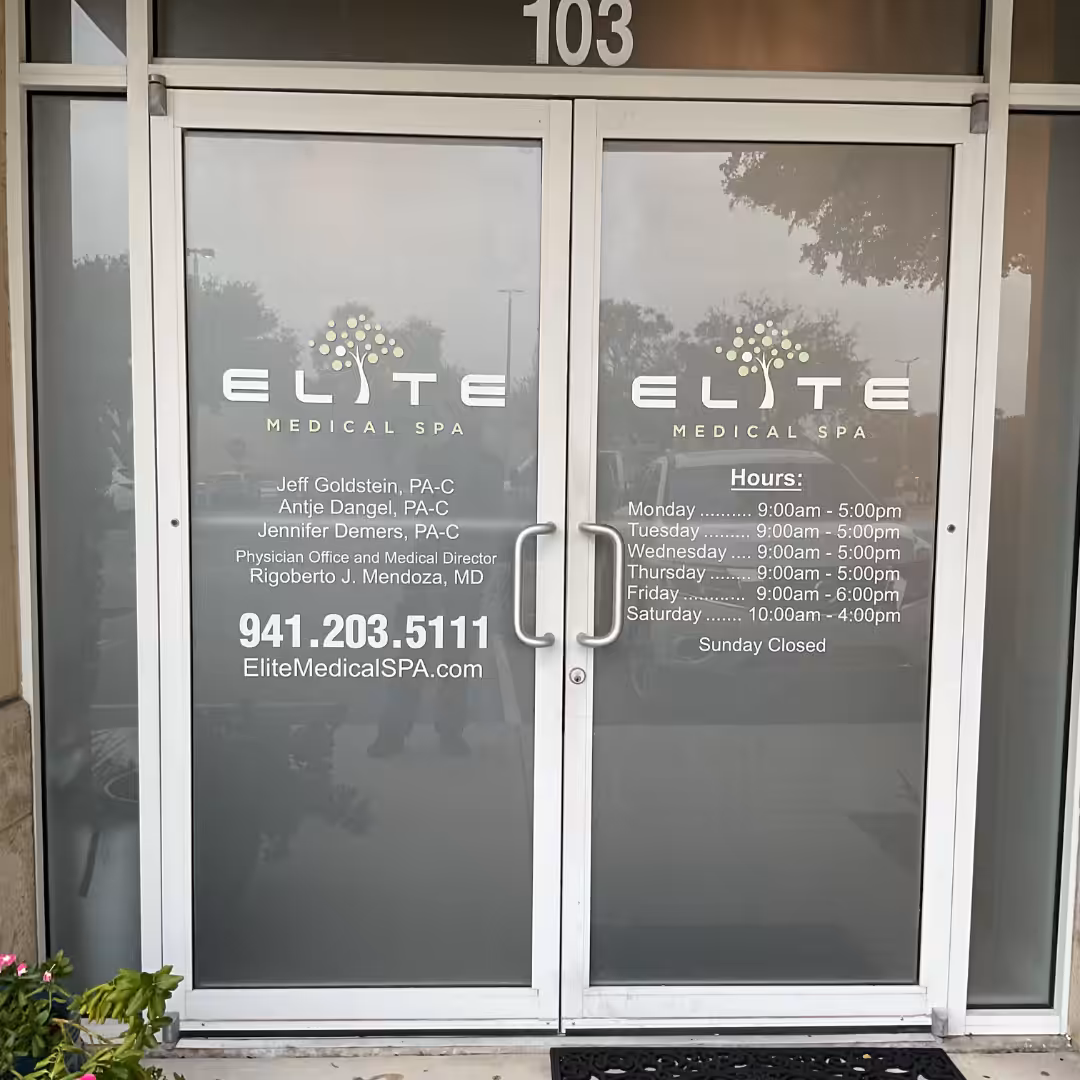 Front doors of Elite Medical Spa feature frosted glass displaying contact info, staff names, website, business hours, and healthcare details. The spa’s logo with green and yellow dots is displayed above the text.
