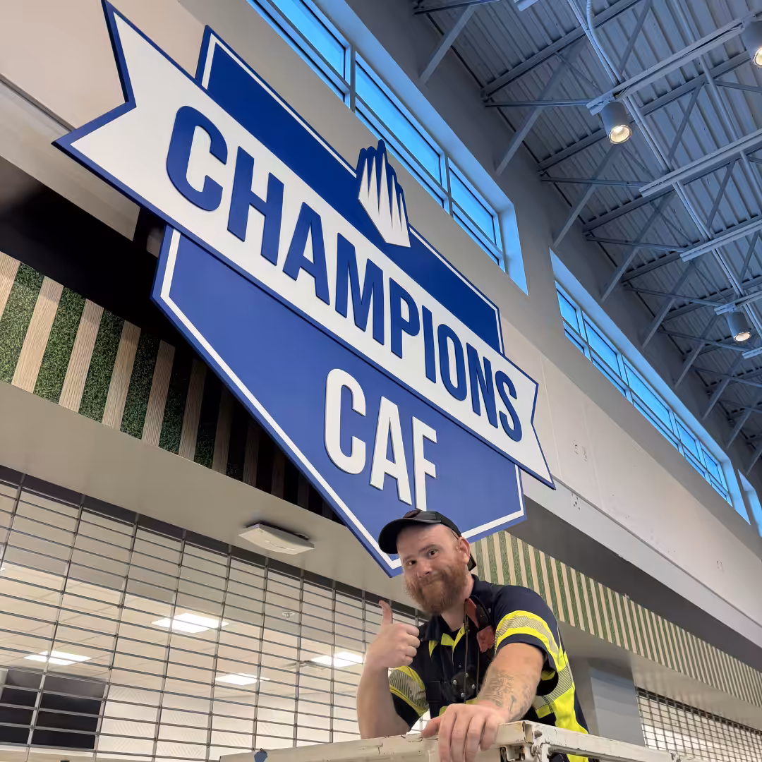 A man in a work uniform gives a thumbs up under a large blue and white CHAMPIONS CAF sign inside a high-ceilinged building in Lakewood Ranch.