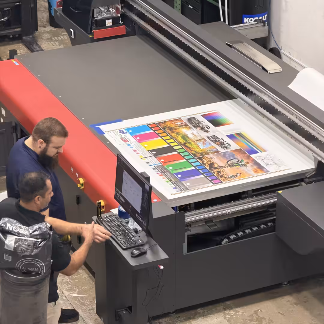 Two men operate a large flatbed printer in Venice, examining a color test print with graphics and color swatches. One man uses the computer, while the other observes the output on the printer’s surface.