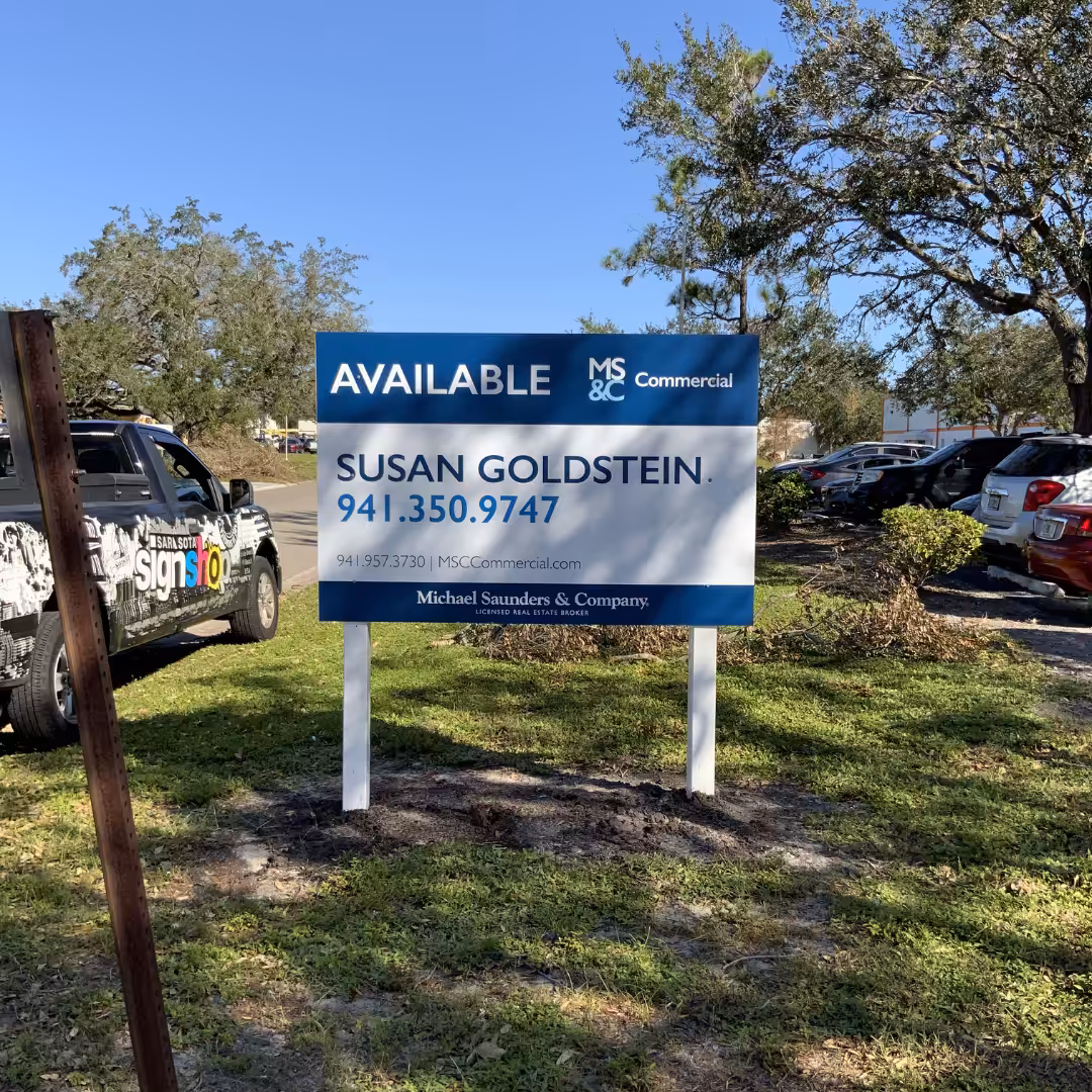 A real estate sign in a parking lot reads Available with contact info for Susan Goldstein from Michael Saunders & Company, surrounded by parked cars and trees in the background.