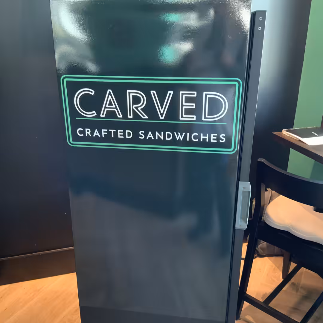 A black refrigerator with a neon green sign that reads CARVED CRAFTED SANDWICHES stands next to a black chair and a table with papers, creating a warm hospitality vibe in the softly lit room.