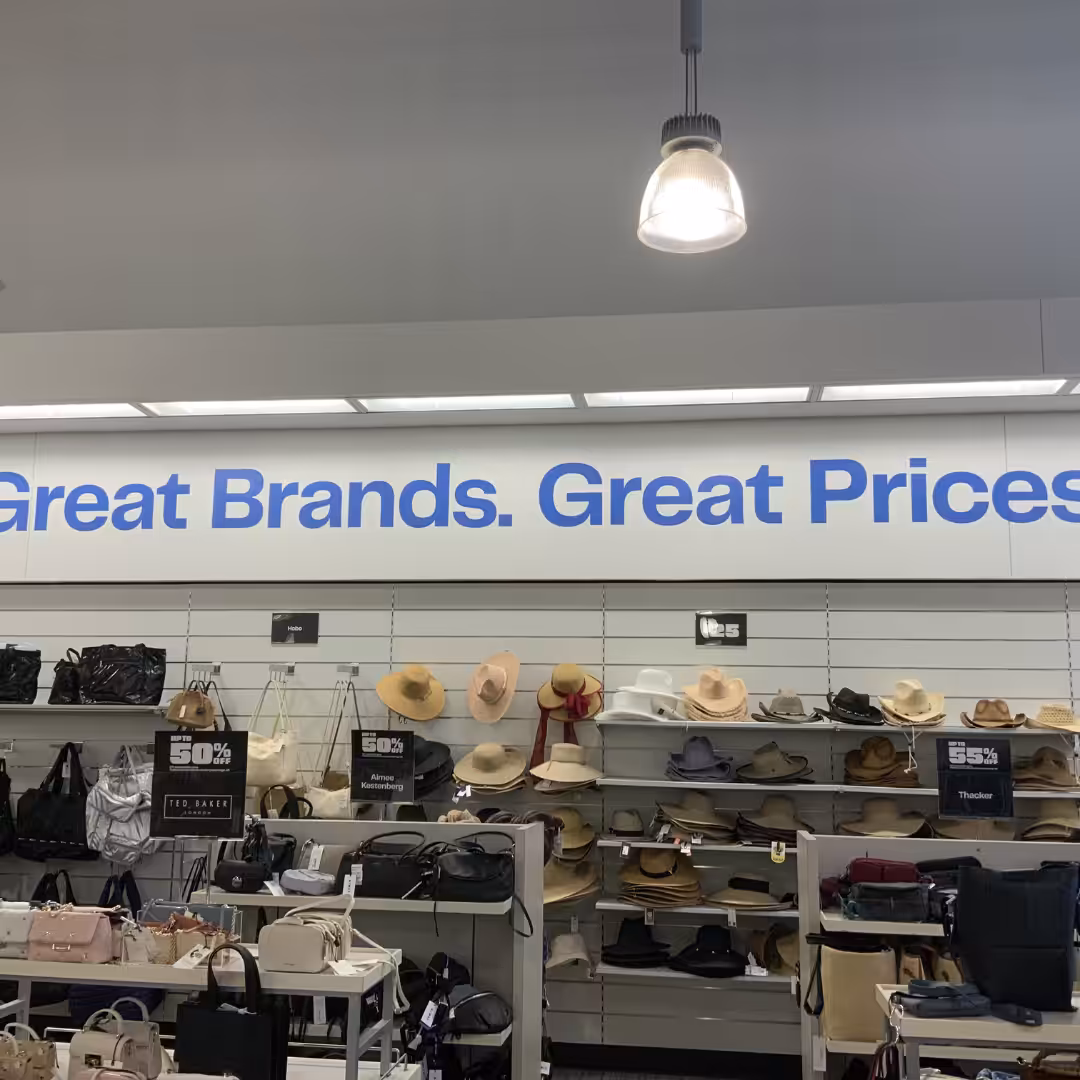 A retail store display features hats, handbags, and purses below a wall sign that reads Great Brands. Great Prices. A sale sign offering 50% off is visible among the merchandise.