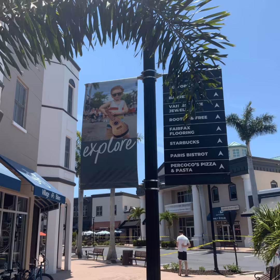Shopping plaza with palm trees, a sign listing nearby stores, and a large banner showing a young child with sunglasses and a guitar under the word explore. Builders & Developers have created an inviting space under blue skies.