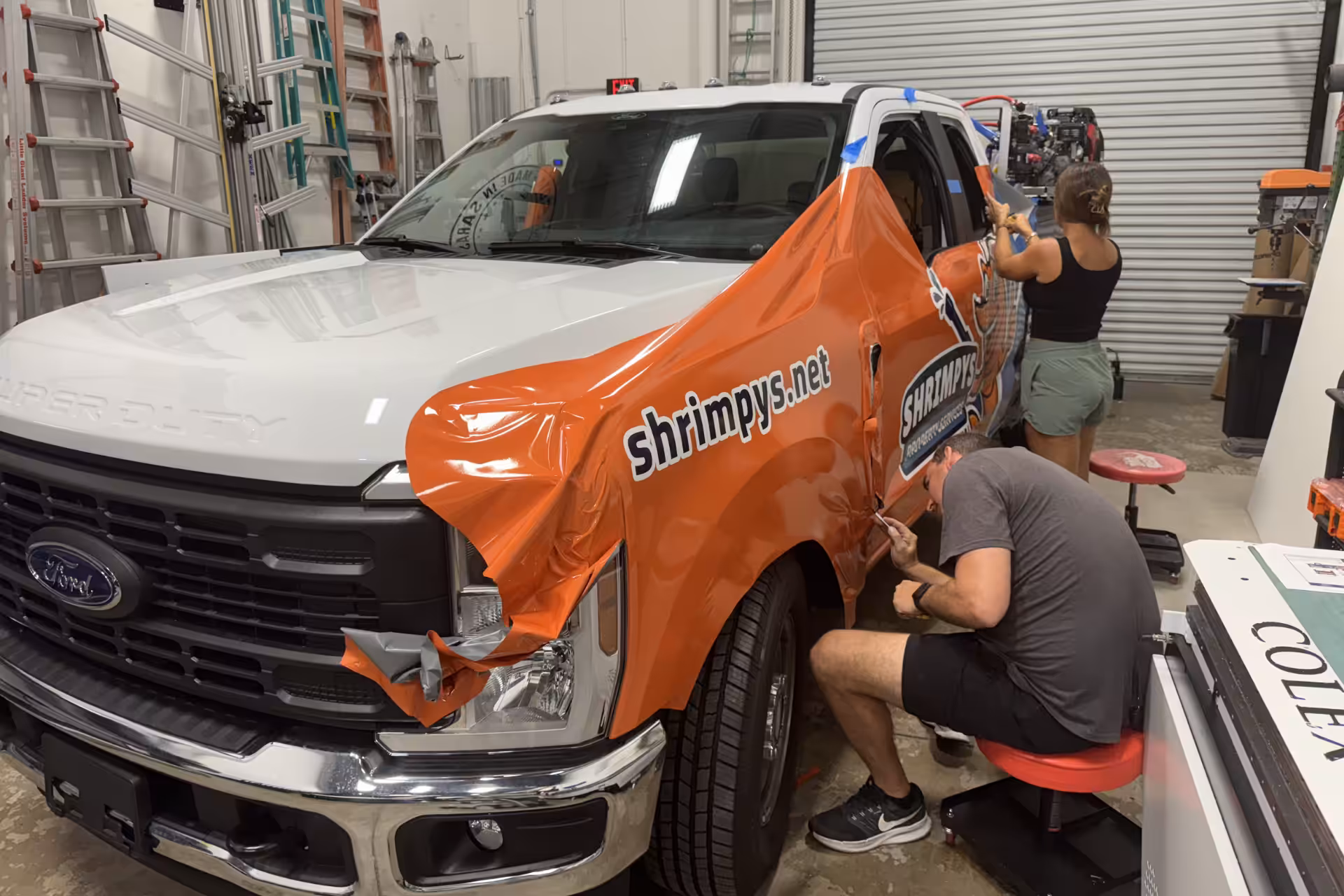 Two people apply an orange wrap with shrimpys.net text and logo to the side of a white Ford truck inside a Sarasota workshop, with tools and equipment visible in the background.