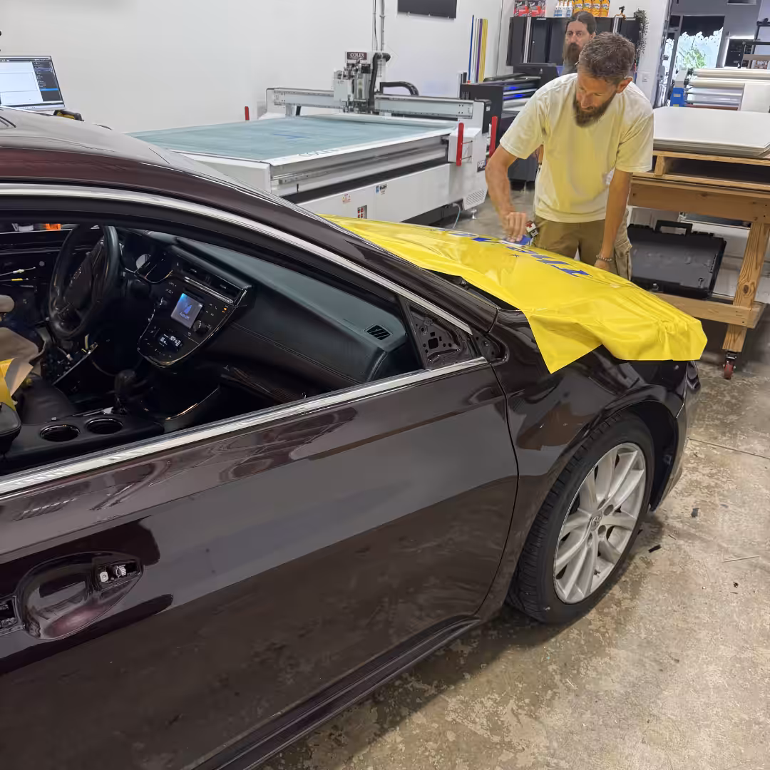 A man at a Lakewood Ranch workshop applies a sheet of yellow vinyl to the front fender of a dark sedan, preparing for a car wrap.