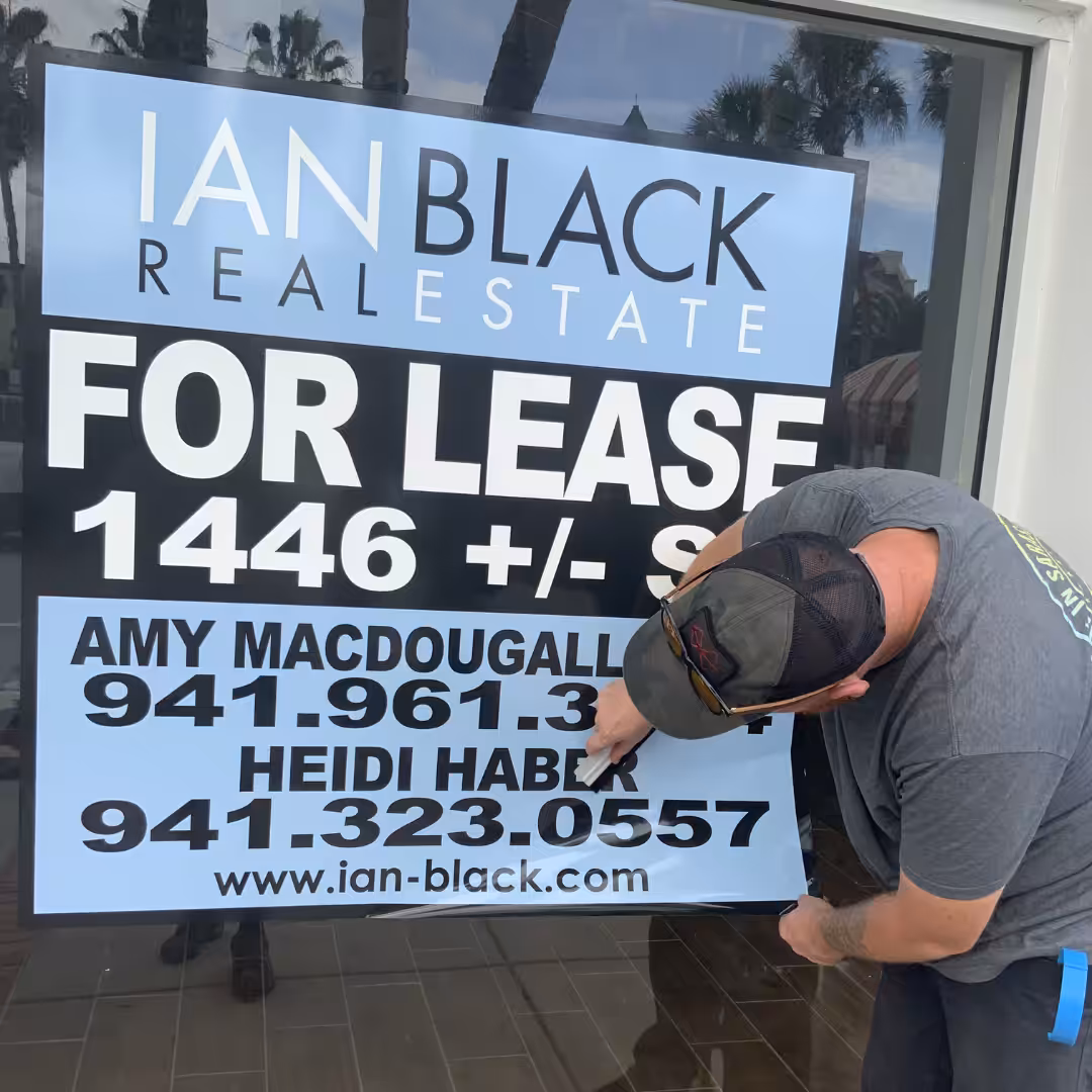 A man points at a large For Lease sign on a Venice storefront window, showing contact information for real estate agents and a website address. Palm trees are reflected in the glass.