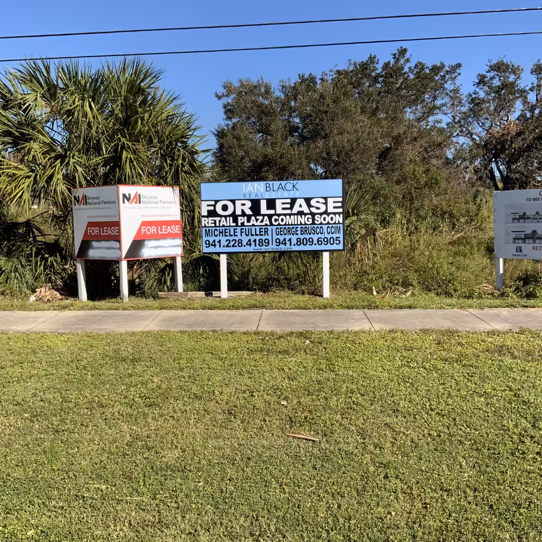 Three For Lease signs stand by a sidewalk in front of bushes and trees, advertising available real estate plaza rental space with contact numbers. The sky is clear and blue.