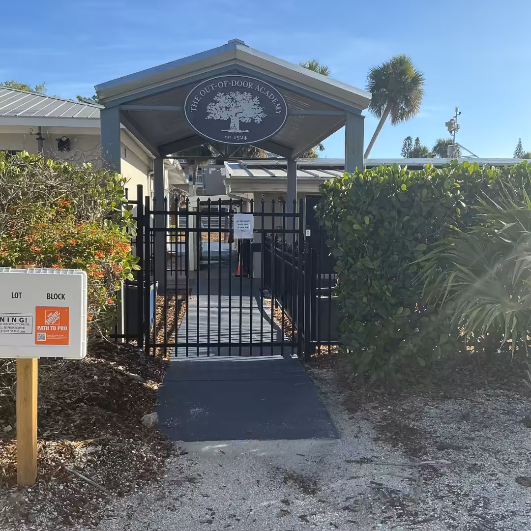 Gated entrance to The Out-of-Door Academy, a leader in schools & sports, with a ramp leading to the door, lush greenery, and a sign for parking information on the left.