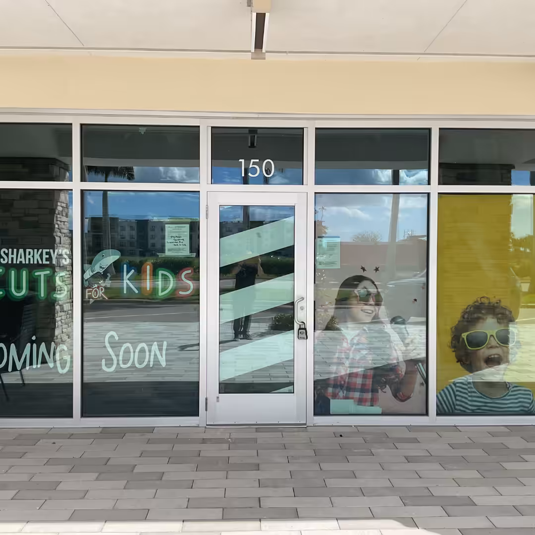 Storefront with windows showing Sharkeys Cuts for Kids Coming Soon and posters of two smiling children, one with sunglasses, indicating a future kids haircut retail salon at suite 150.