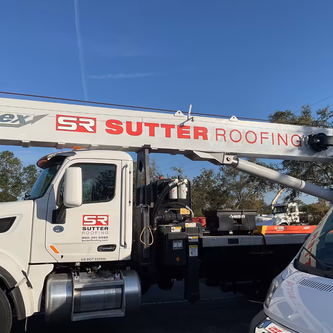 A white Home Service truck with a large crane arm labeled Sutter Roofing is parked outdoors on a sunny day, with trees and a blue sky in the background. The truck door displays the Sutter Roofing logo and contact information.