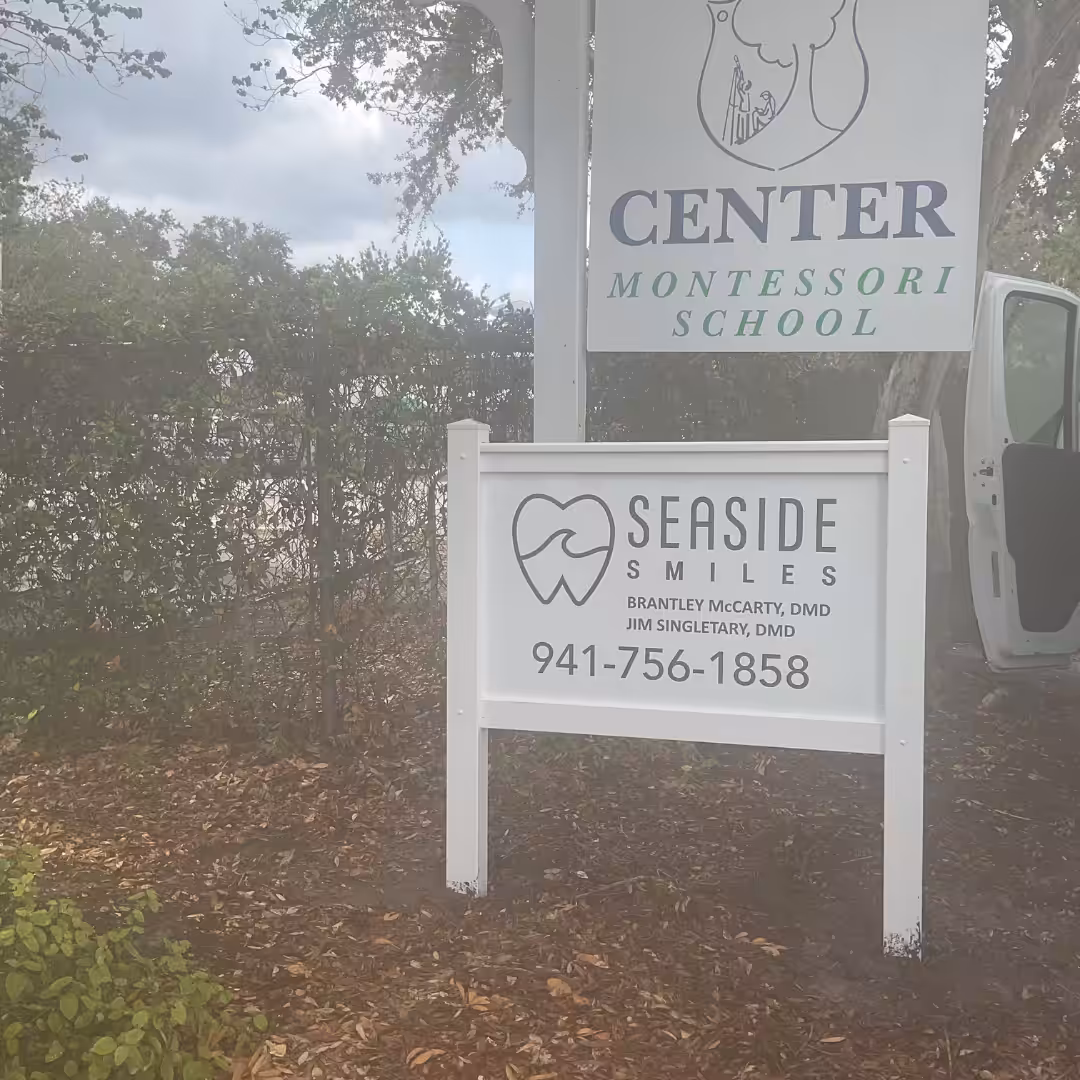 White sign for Seaside Smiles dental office with phone number stands in front of a Montessori school sign, highlighting the connection between schools & sports. Bushes, trees, and an open car door are visible in the background.