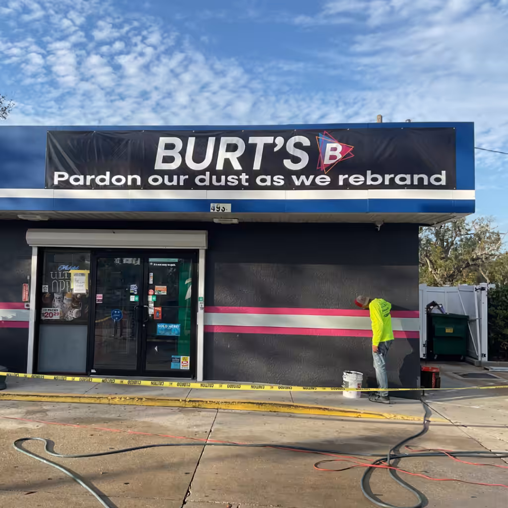 A worker in a yellow hoodie paints the exterior wall of BURTS, with banners and a sign above reading, Pardon our dust as we rebrand. Caution tape and paint supplies are scattered on the ground.