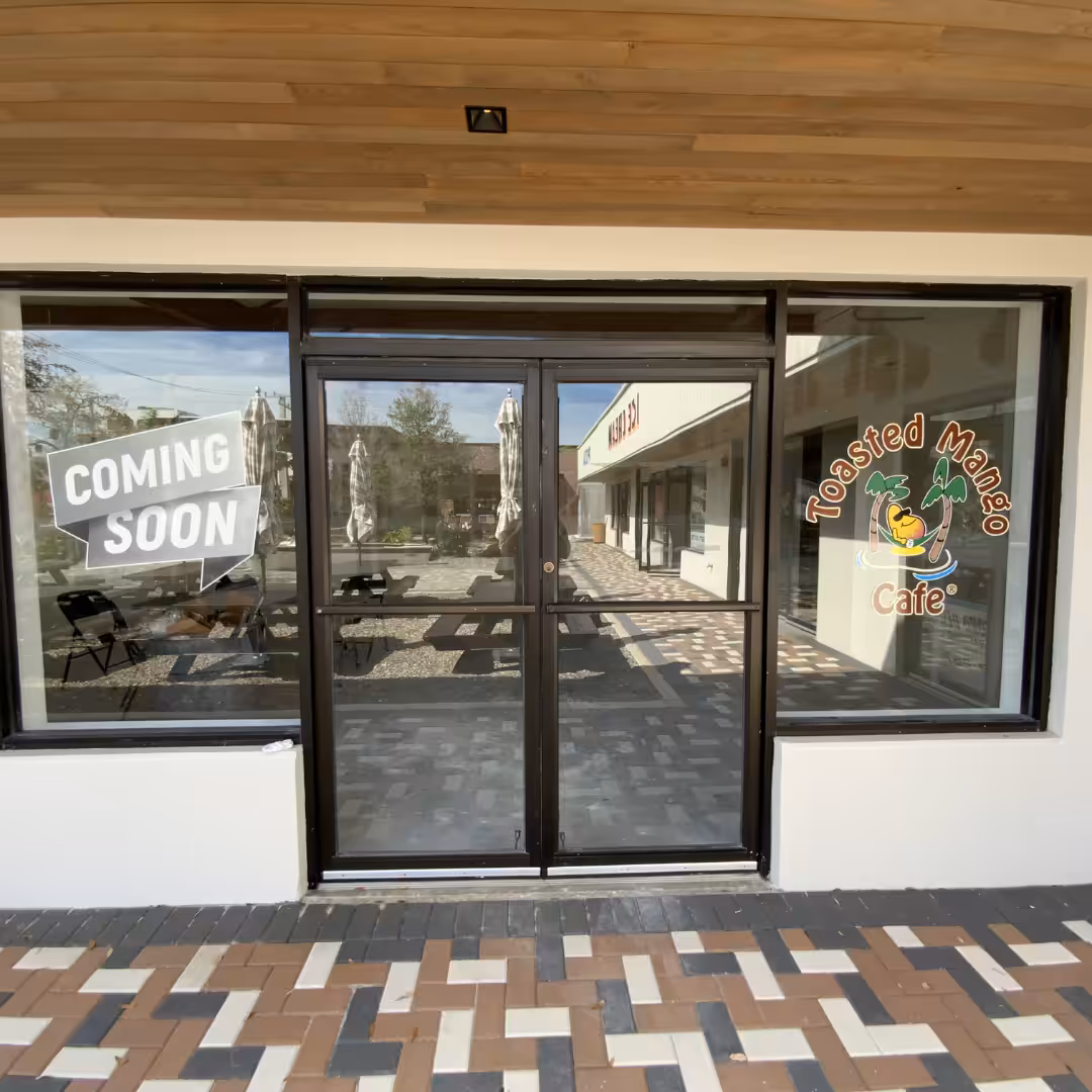 Glass storefront with COMING SOON sign in one window and a Toasted Mango Café logo on the other, reflecting an inviting outdoor patio. A brick-patterned walkway welcomes guests to experience true hospitality.