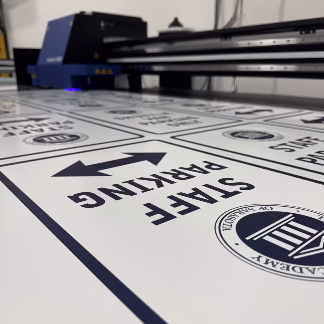 Close-up of a large printer producing multiple STAFF PARKING signs with arrows, an official emblem, and designs suited for schools & sports facilities. Both the printer and finished signs are clearly visible.