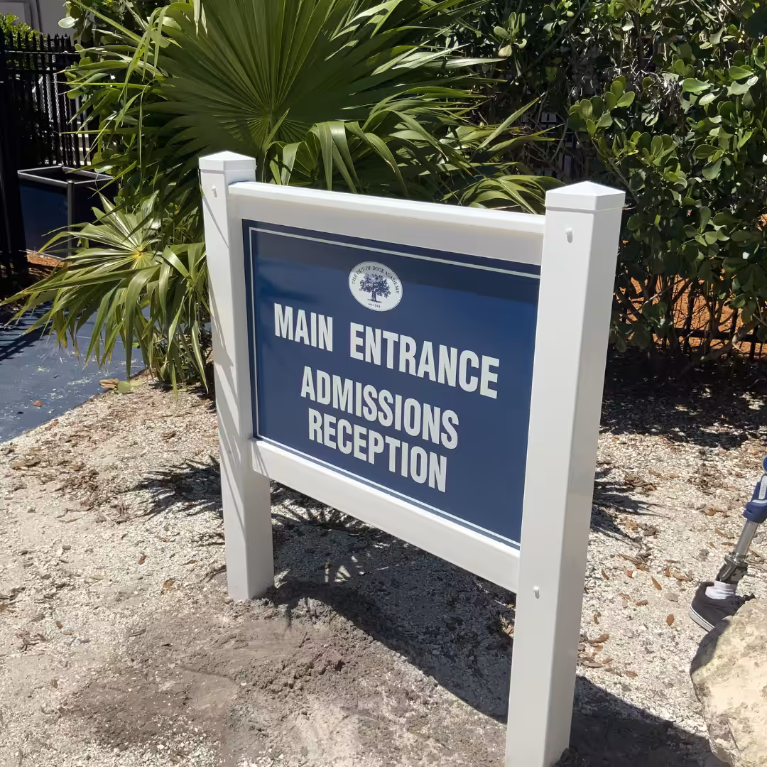 A blue and white outdoor sign reads: MAIN ENTRANCE ADMISSIONS RECEPTION, guiding visitors to schools & sports facilities. It stands on gravel with green plants and palm leaves in the background.