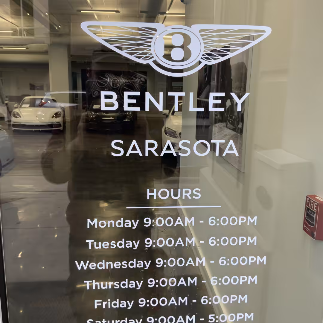 A glass retail door with a Bentley logo and Bentley Sarasota above business hours listed for each day of the week. Luxury cars are visible inside through the glass.