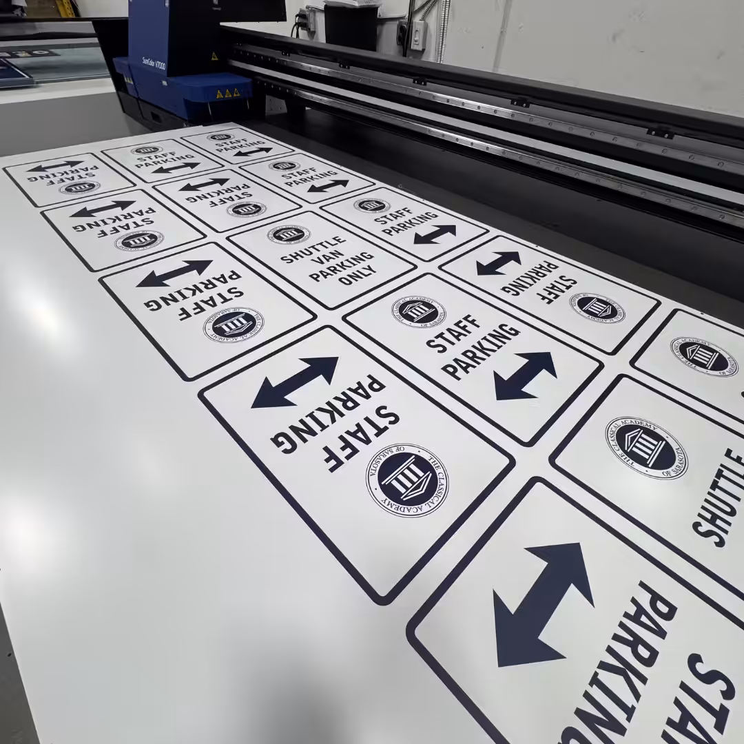 Sheets of printed parking and shuttle signs with arrows and institutional logos—perfect for schools & sports events—are laid out on a surface, likely being prepared by a large-format printer in an industrial setting.