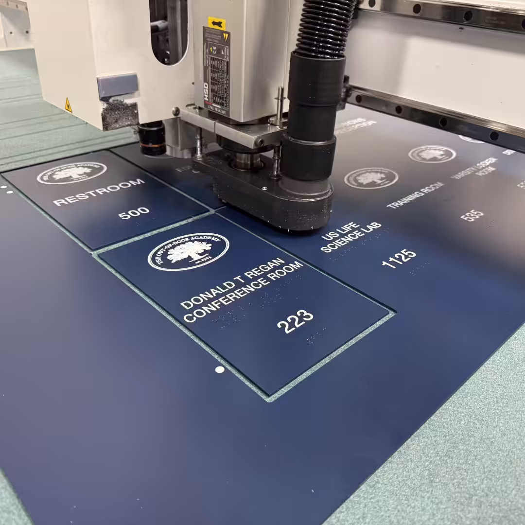 A machine engraves blue signage boards for schools & sports facilities, including labels for restrooms, conference rooms, science labs, and parking access—all with white text and icons.