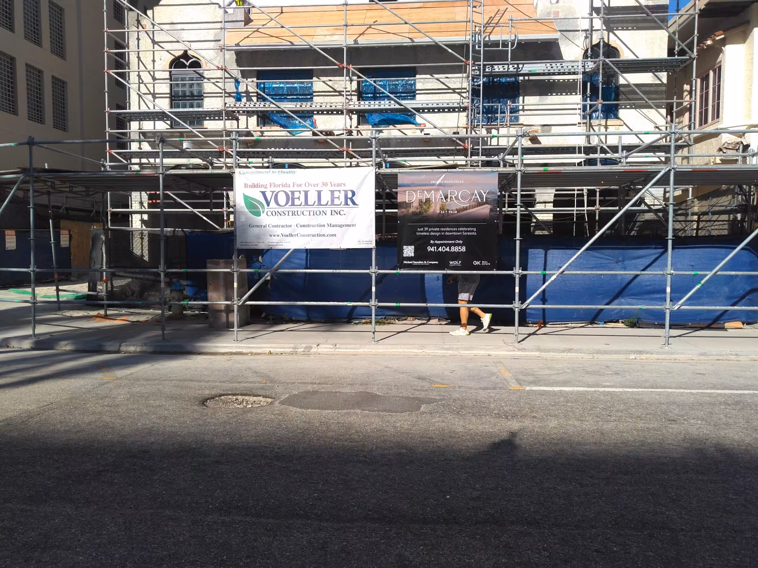 A person walks behind scaffolding in front of a bradenton building under construction. Two signs are visible—one for Voeller Construction Inc. and one for Demarcay. Shadows cover part of the street.