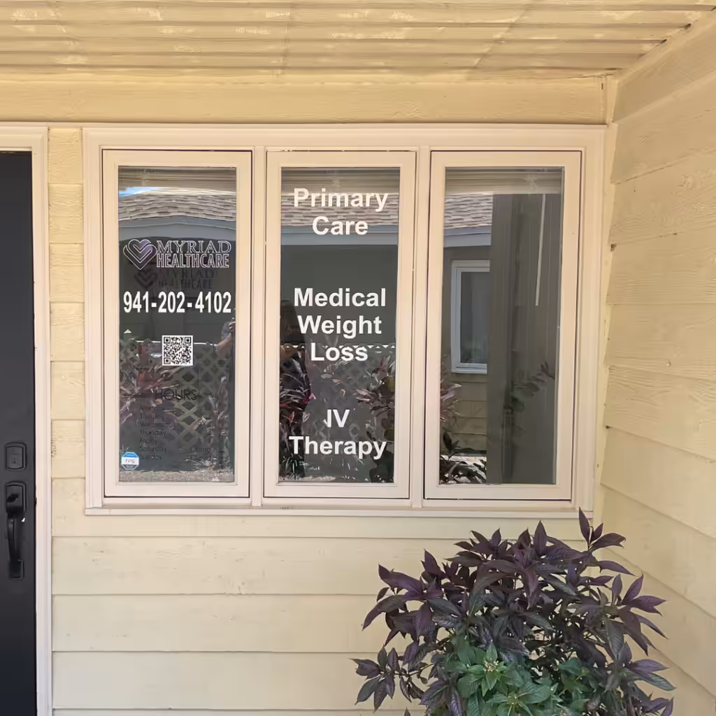 Three windows on a beige building display Sarasota Sign for Mermaid Healthcare, listing Primary Care, Medical Weight Loss, and IV Therapy. A phone number, QR code, and plants are visible in front of the windows.