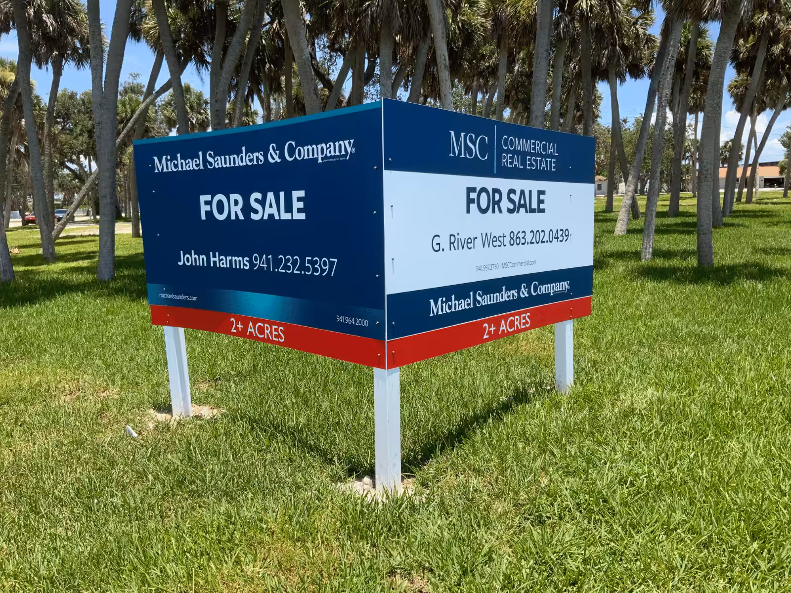 Large blue and white Sarasota Sign for sale by Michael Saunders & Company stands on grass, advertising over 2 acres. Contact numbers for John Harms and G. River West. Palm trees visible in the background.