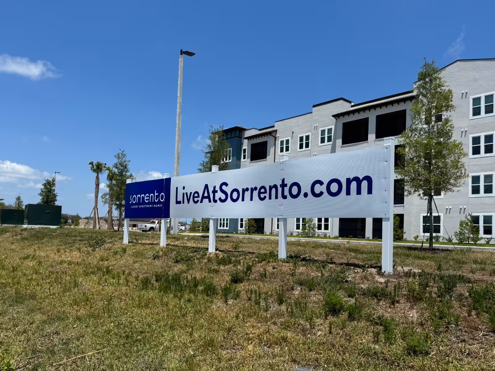 A Sarasota Sign reads LiveAtSorrento.com in front of a modern apartment building under a clear blue sky, with grass and a few trees in the foreground.