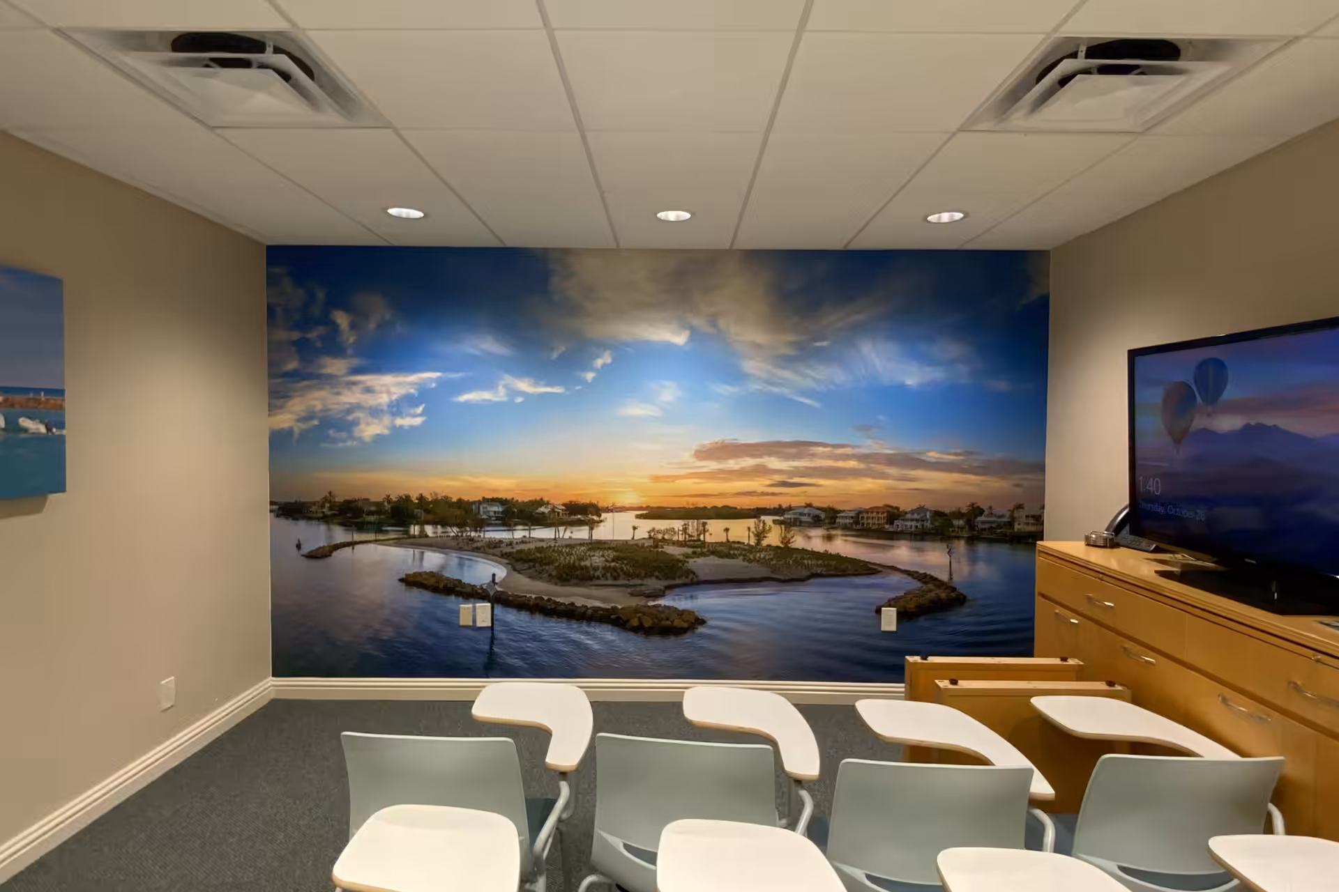 Small Sarasota classroom with white chairs and desks facing a wall mural of a lakeside sunset. A TV stands on a wooden cabinet to the right. Ceiling lights are on, and walls are beige.